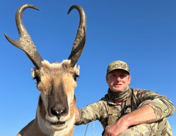 A hunter in full camouflage smiles while kneeling behind a pronghorn antelope. The photo highlights the stunning Wyoming prairie under a clear blue sky.